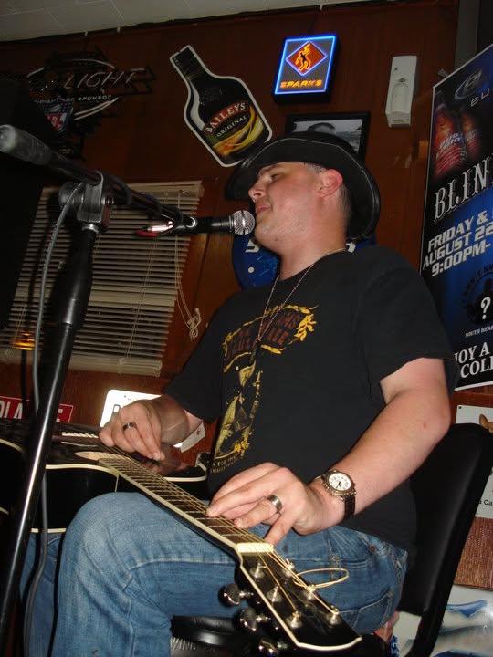Blind Joe in a black cowboy hat playing his flat-top acoustic guitar across his lap at a bar gig, singing into a mic.