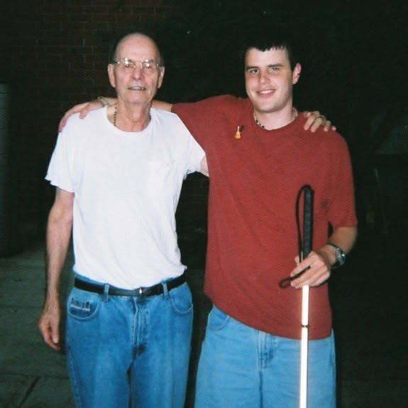 A younger Blind Joe in a red T-shirt and jeans, holding his white cane, standing arm in arm with an older man in a white T-shirt.