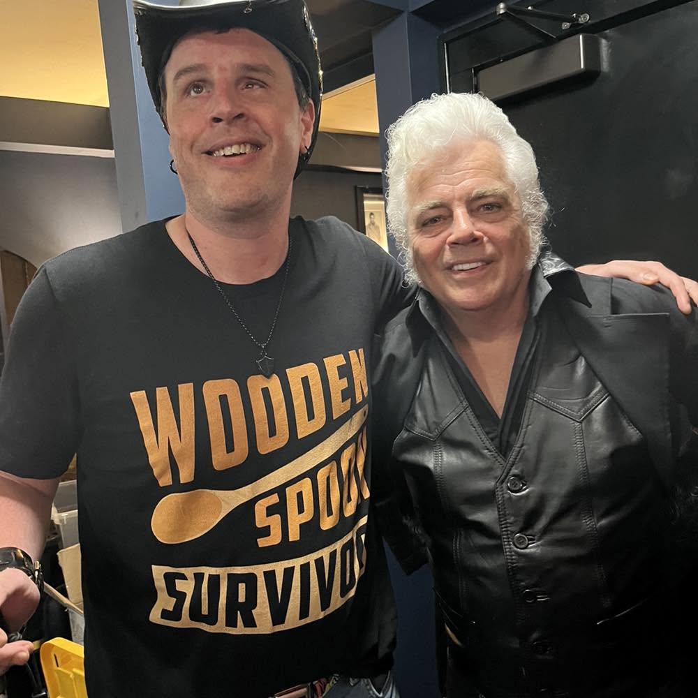Blind Joe backstage in a black cowboy hat and a black Wooden Spoon Survivor T-shirt, smiling with his arm around a man with snow-white hair in a black leather jacket.
