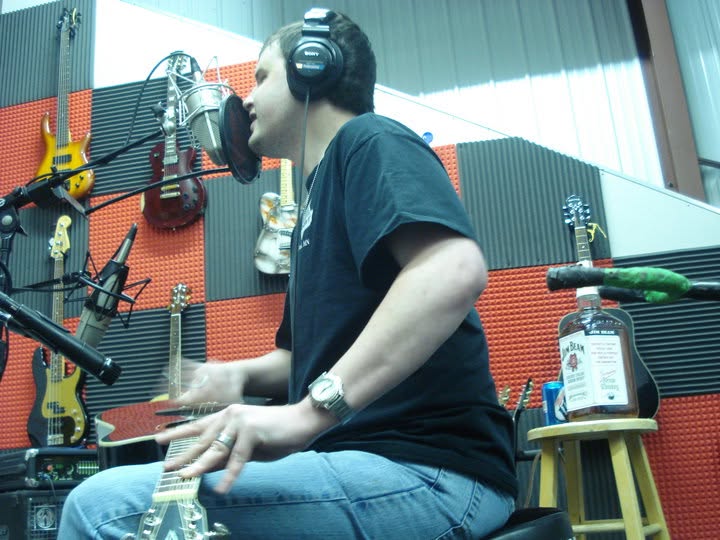 Blind Joe in a recording studio wearing headphones, singing into a big studio microphone with guitars hanging on the wall behind him.