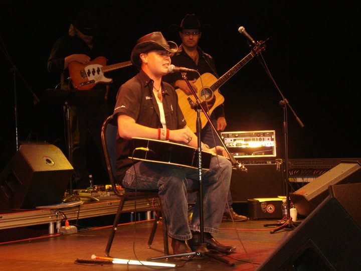 Blind Joe sitting on a stage under warm lights, playing an acoustic guitar and singing into a mic.