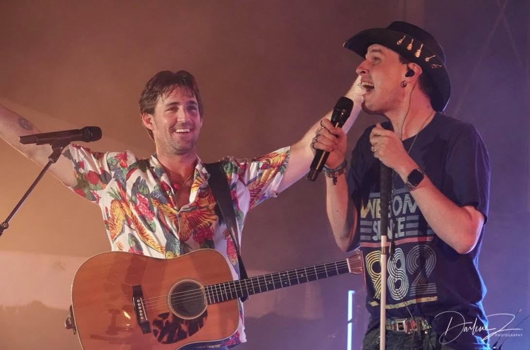 Blind Joe in a black cowboy hat singing into a handheld mic on stage, side by side with a singer in a bright Hawaiian shirt who's playing an acoustic guitar, under warm purple stage lights.
