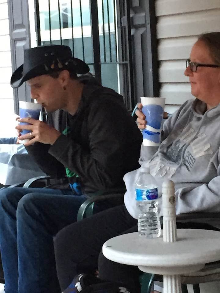 Blind Joe in a black cowboy hat and black jacket, sipping from a coffee cup on a front porch beside his wife Liann, who has glasses and a gray sweatshirt and is sipping from her own cup.