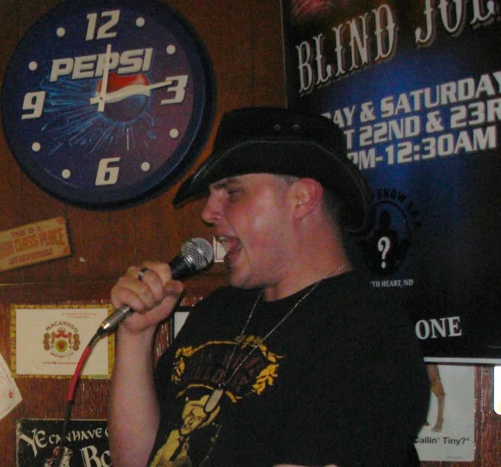Blind Joe in a black cowboy hat belting into a microphone in a bar, with a Pepsi clock and a sign for his weekend shows on the wall behind him.