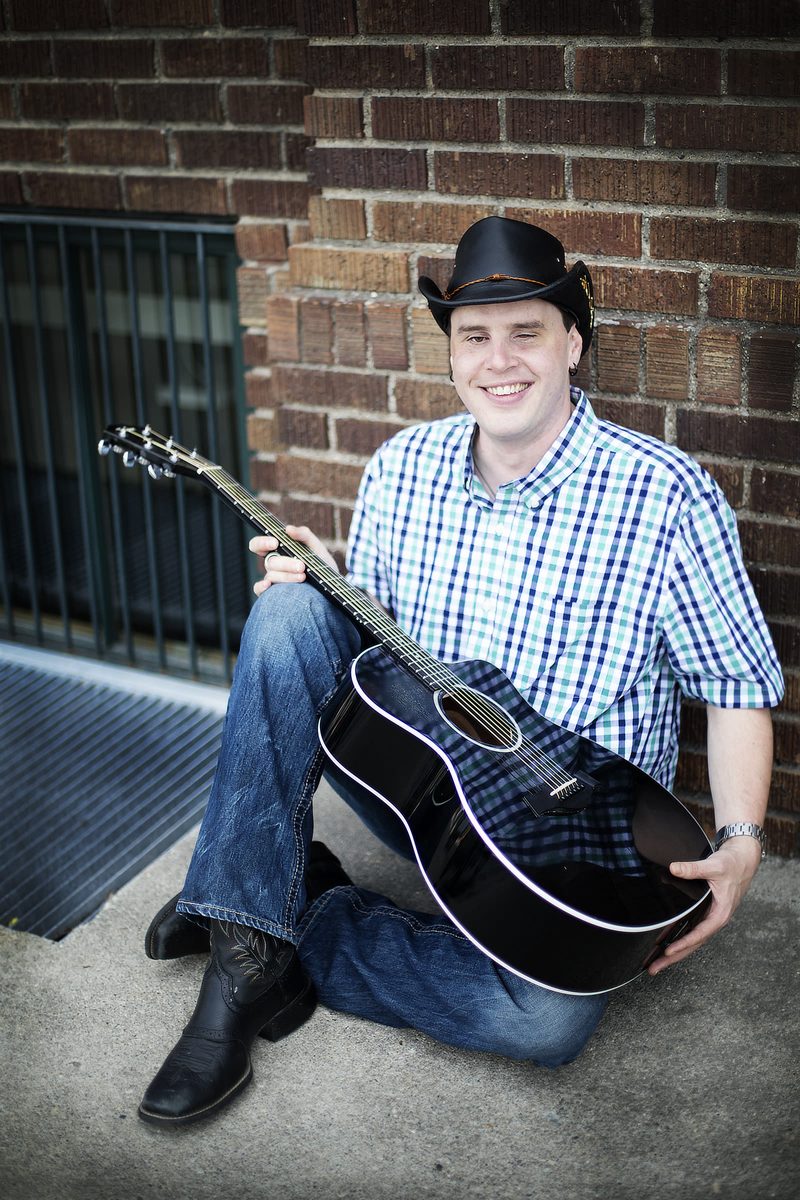 Blind Joe sitting on the sidewalk against a red brick wall, smiling, in a black cowboy hat and plaid shirt, holding his black acoustic guitar across his lap.