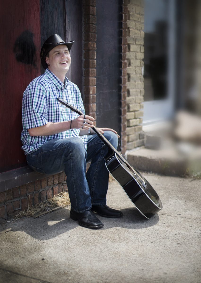 Blind Joe laughing, sitting on a low brick ledge in front of an old red wooden door, with his black guitar leaning against his leg.