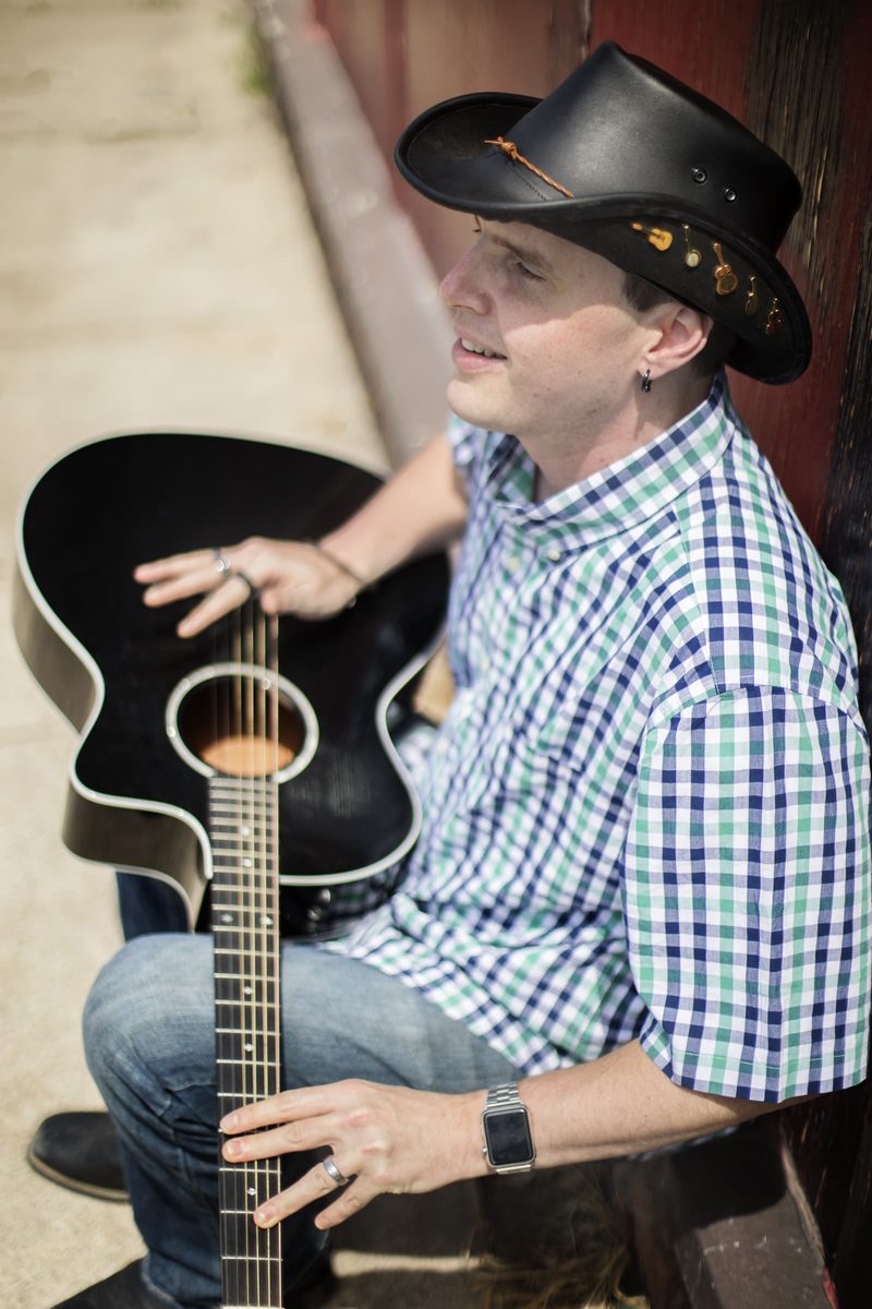 Close-up of Blind Joe from above, smiling with his hands on his black acoustic guitar. Little guitar pins line the band of his black cowboy hat.