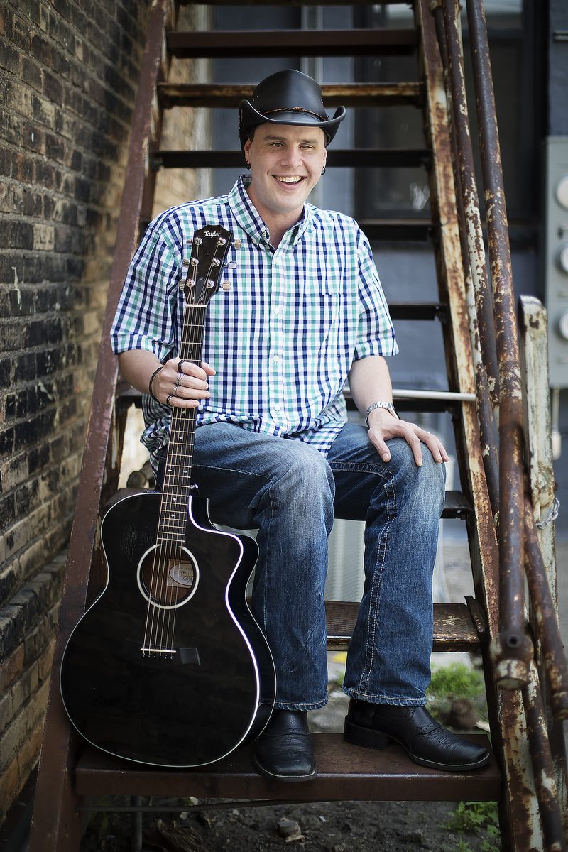 Blind Joe grinning on an old rusty metal fire escape, wearing a black cowboy hat, a blue and green plaid shirt, jeans and black boots, with his black acoustic guitar standing beside him.