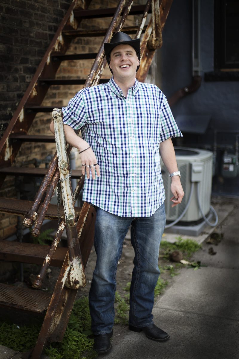 Blind Joe laughing, leaning on the railing of a rusty fire escape in an alley, in his black cowboy hat, plaid shirt and jeans.
