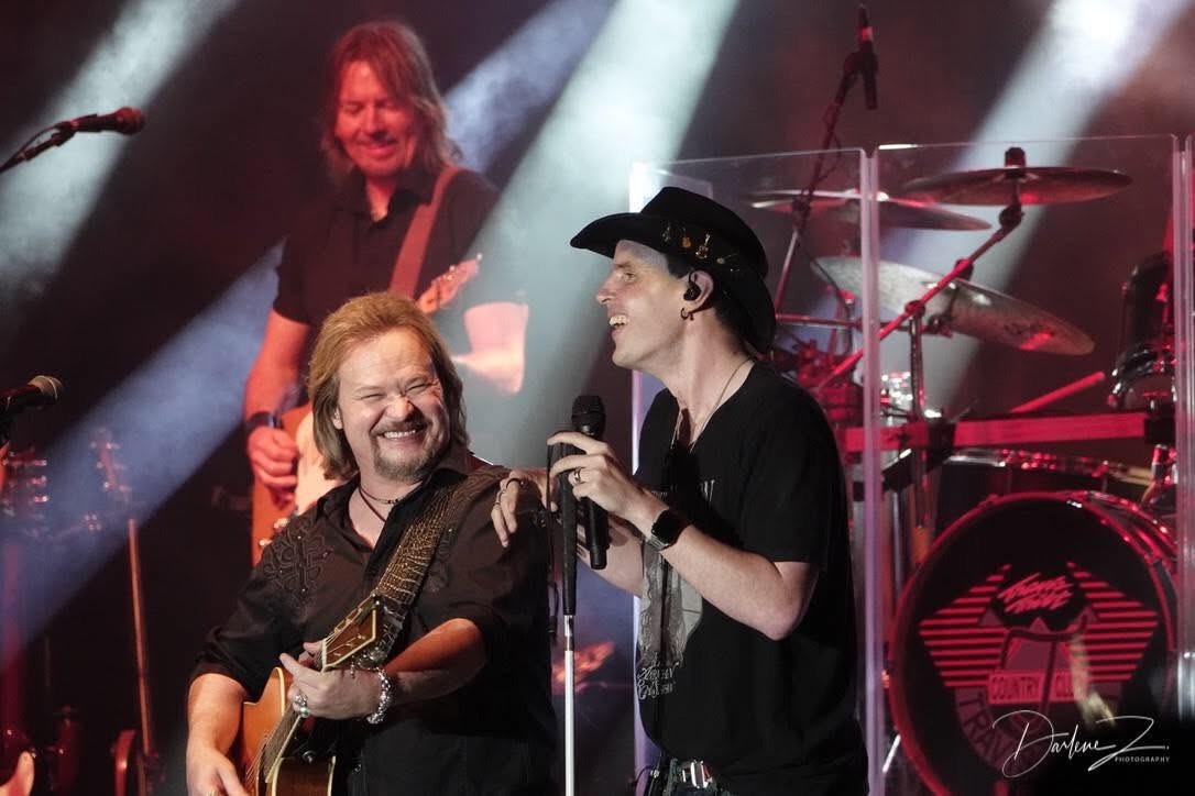 Blind Joe in his black cowboy hat and black T-shirt, singing into a headset mic on a concert stage beside Travis Tritt, who's playing guitar and grinning. Bright white stage lights and a drummer behind them.