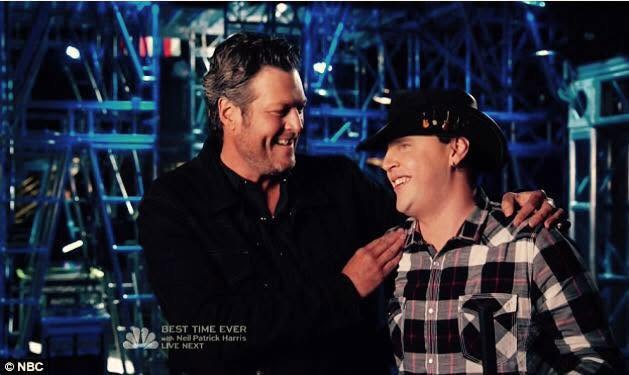 Blind Joe in a black cowboy hat and plaid shirt, laughing with his coach Blake Shelton, who has his arm around him on the set of The Voice.