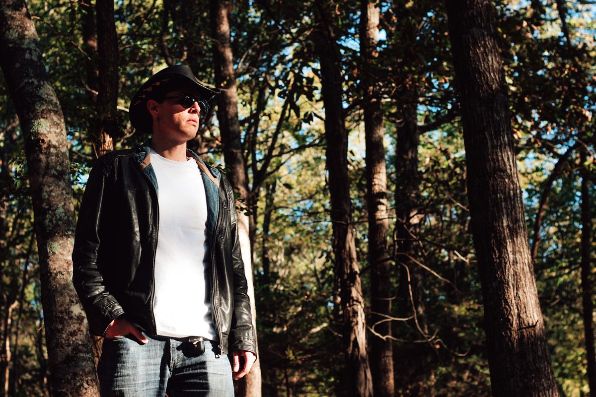 Blind Joe in a black cowboy hat, sunglasses, black leather jacket, white T-shirt and jeans, standing among tall trees in late-afternoon sun.